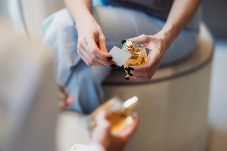 A close-up of a woman holding a scent strip and an elegant perfume bottle, showcasing fragrance testing in a well-lit environment. The setting emphasizes the sensory experience and art of perfumery.