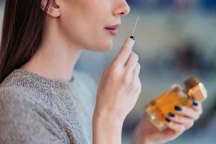 A close-up of a woman holding a scent strip and an elegant perfume bottle, showcasing fragrance testing in a well-lit environment. The setting emphasizes the sensory experience and art of perfumery.